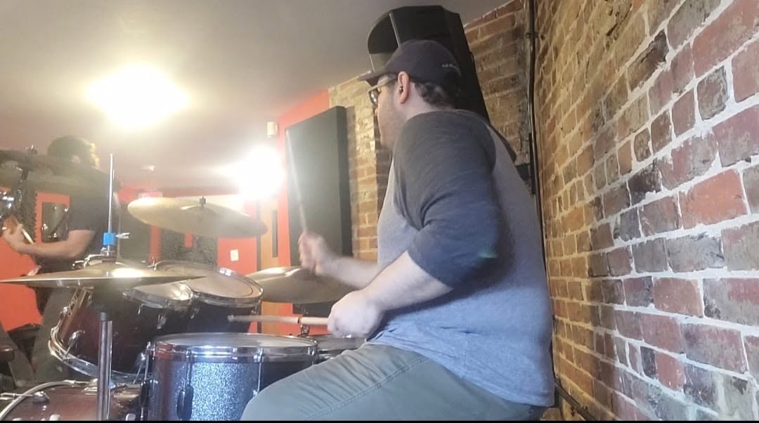 Peter Rodriguez playing drums during a rehearsal beside a brick wall